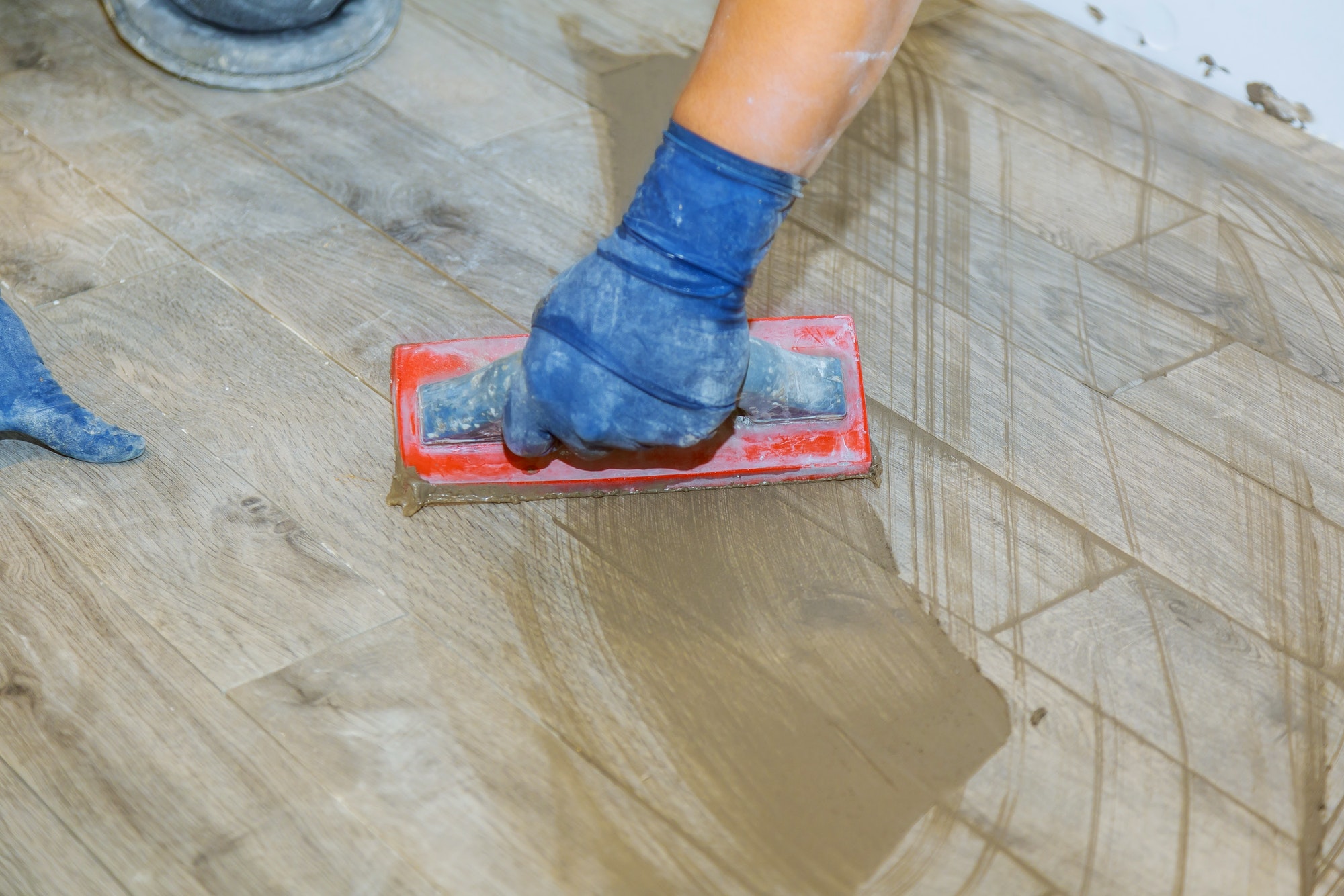 A person in gloves uses a red trowel to smooth a layer of cement on a wooden floor, showcasing a construction project.