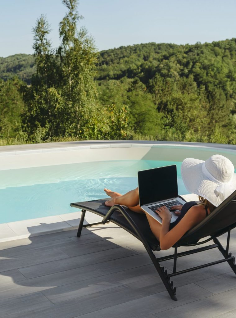 A person relaxes on a lounge chair by a pool, using a laptop, surrounded by green hills and trees under a clear sky. A person relaxes on a lounge chair by a pool, using a laptop, surrounded by green hills and trees under a clear sky.
